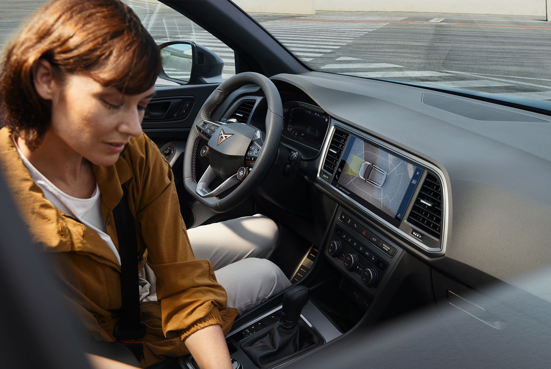 Interior del CUPRA Ateca ressaltant les característiques tecnològiques avançades. El tauler mostra una gran pantalla d'infotainment amb navegació i el volant amb botons per satèl·lit té controls integrats. Una dona està asseguda a l'interior.