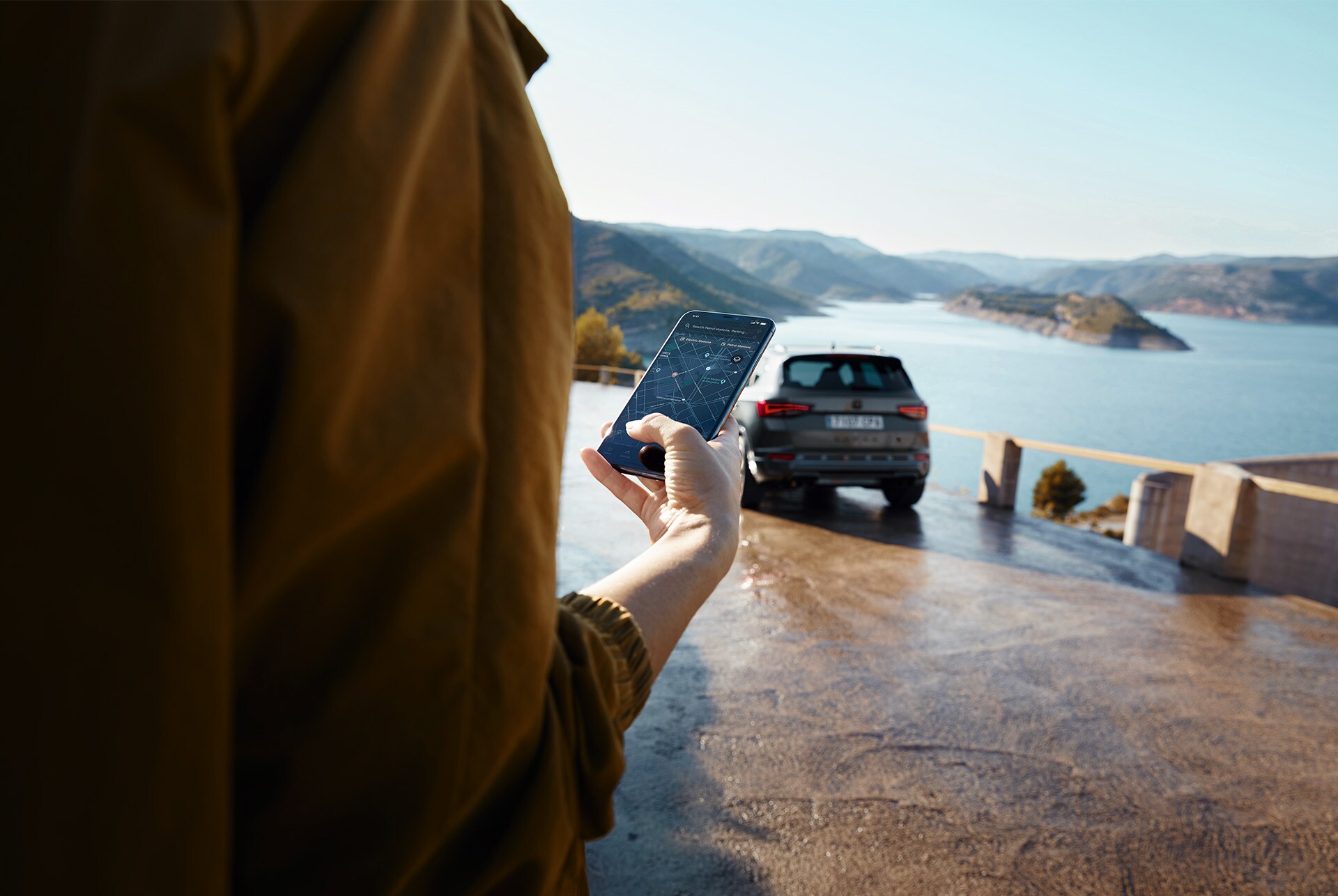 Una persona subjectant un smartphone amb el CUPRA Ateca al fons, estacionat prop d’una vista panoràmica de muntanyes i aigua, simbolitzant un món connectat.