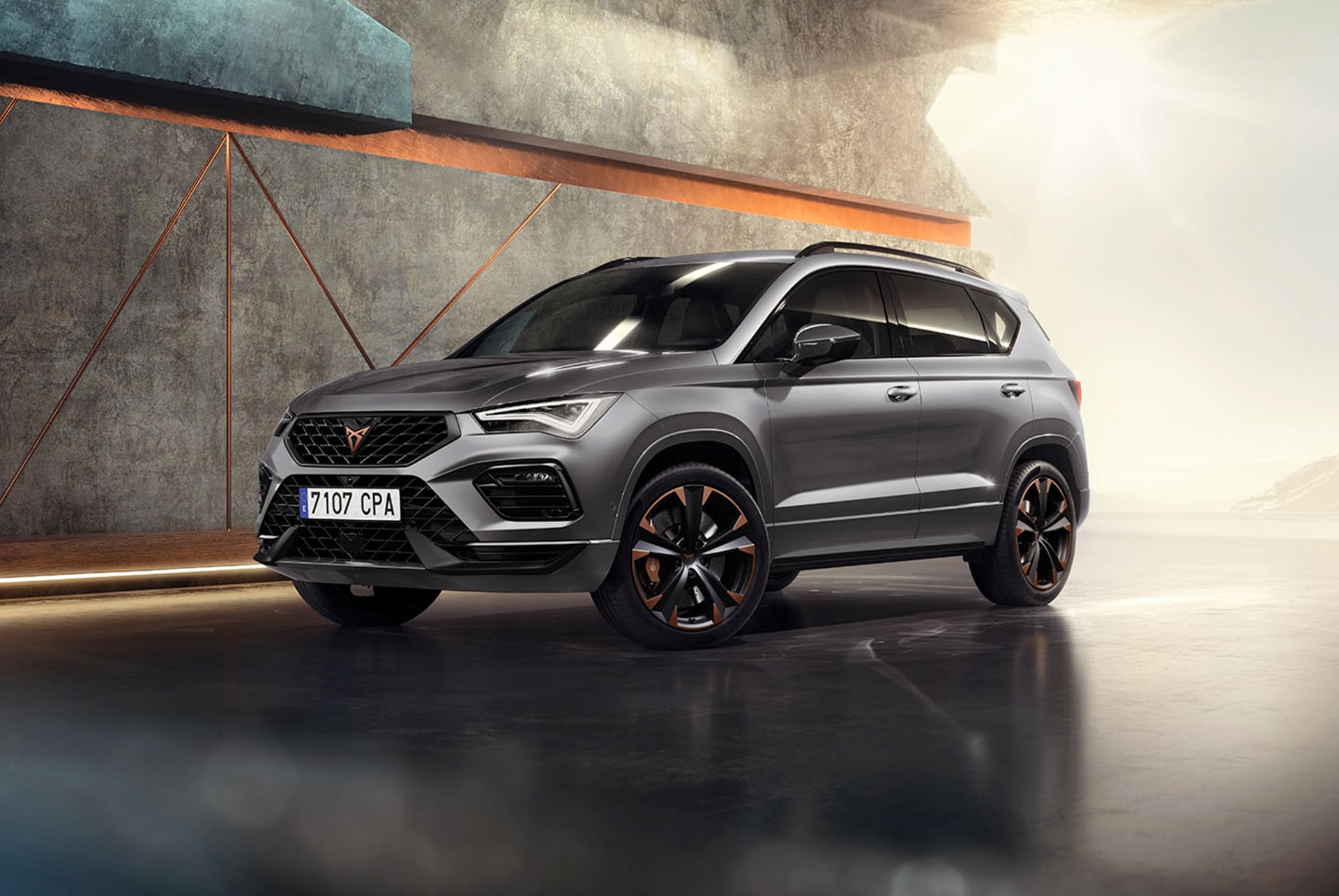 Un CUPRA Ateca 2025 en color Gris Graphene, un SUV esportiu i compacte aparcat en un entorn modern i industrial. El seu disseny destaca per les línies aerodinàmiques, els fars LED i les exclusives llantes d’aliatge amb detalls en coure. Al fons, una paret amb un disseny geomètric contrasta amb les línies elegants del vehicle.