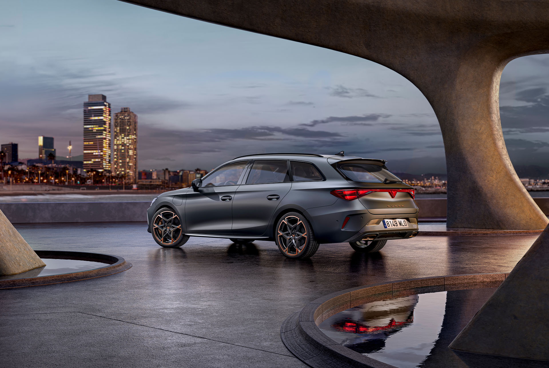 Vista lateral posterior del nou CUPRA Leon Sportstourer 2025, esportiu i aerodinàmic de color gris mat, aparcat en un passeig marítim durant el capvespre. L'skyline de la ciutat brilla de fons.