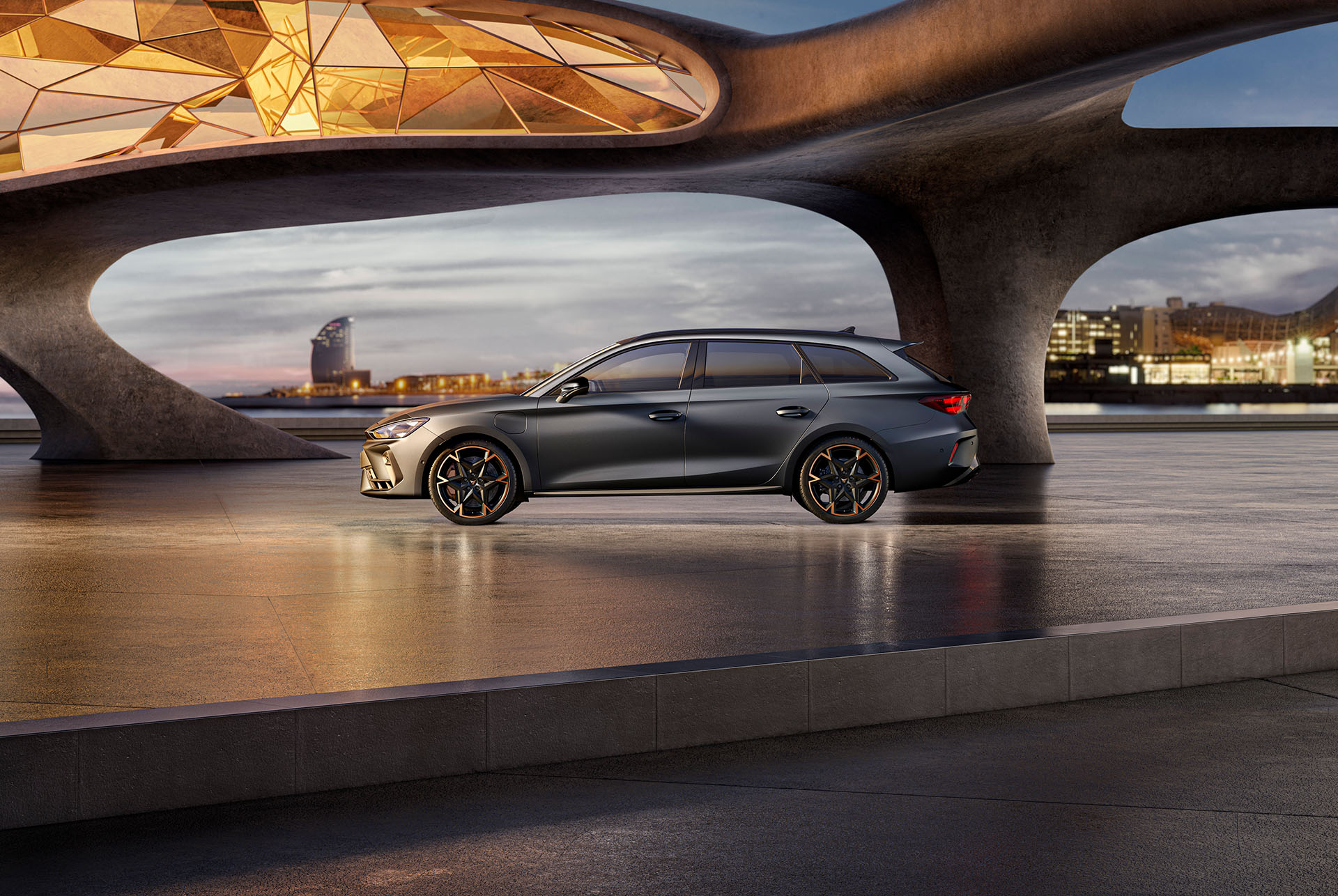 El nou Esportiu Cupra Leon Sportstourer 2025 aparcat sobre formigó, ciutat il·luminada al capvespre en el fons. El vehicle apareix en un acabat gris mat.