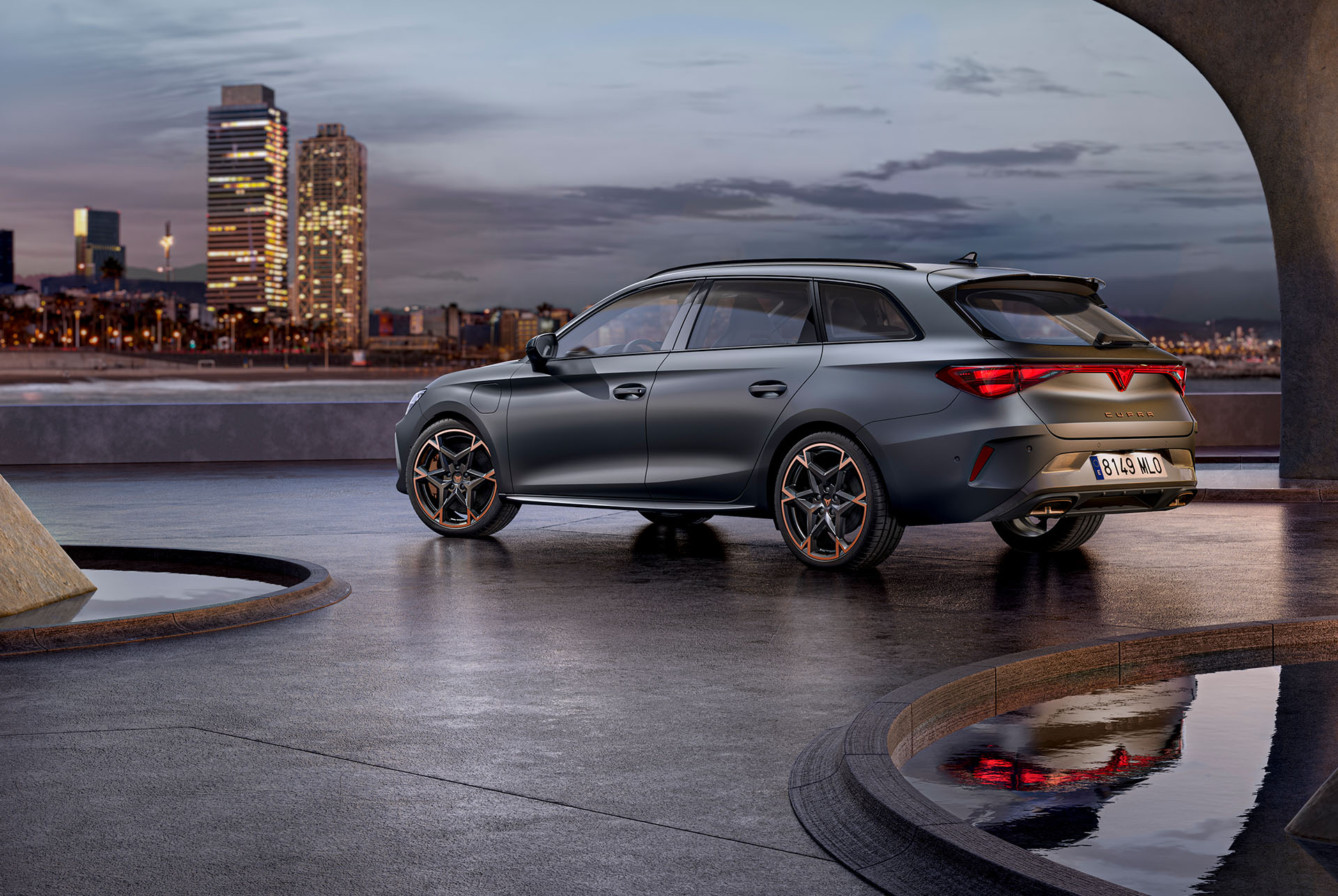 Tecnologia del nou Cupra Leon Sportstourer 2025 aparcat sobre ciment, estructura amb vores brillants, amb vista a l'horitzó de la ciutat al capvespre. El vehicle es mostra amb un acabat gris mat amb llandes d'aliatge en color cobri i negre.