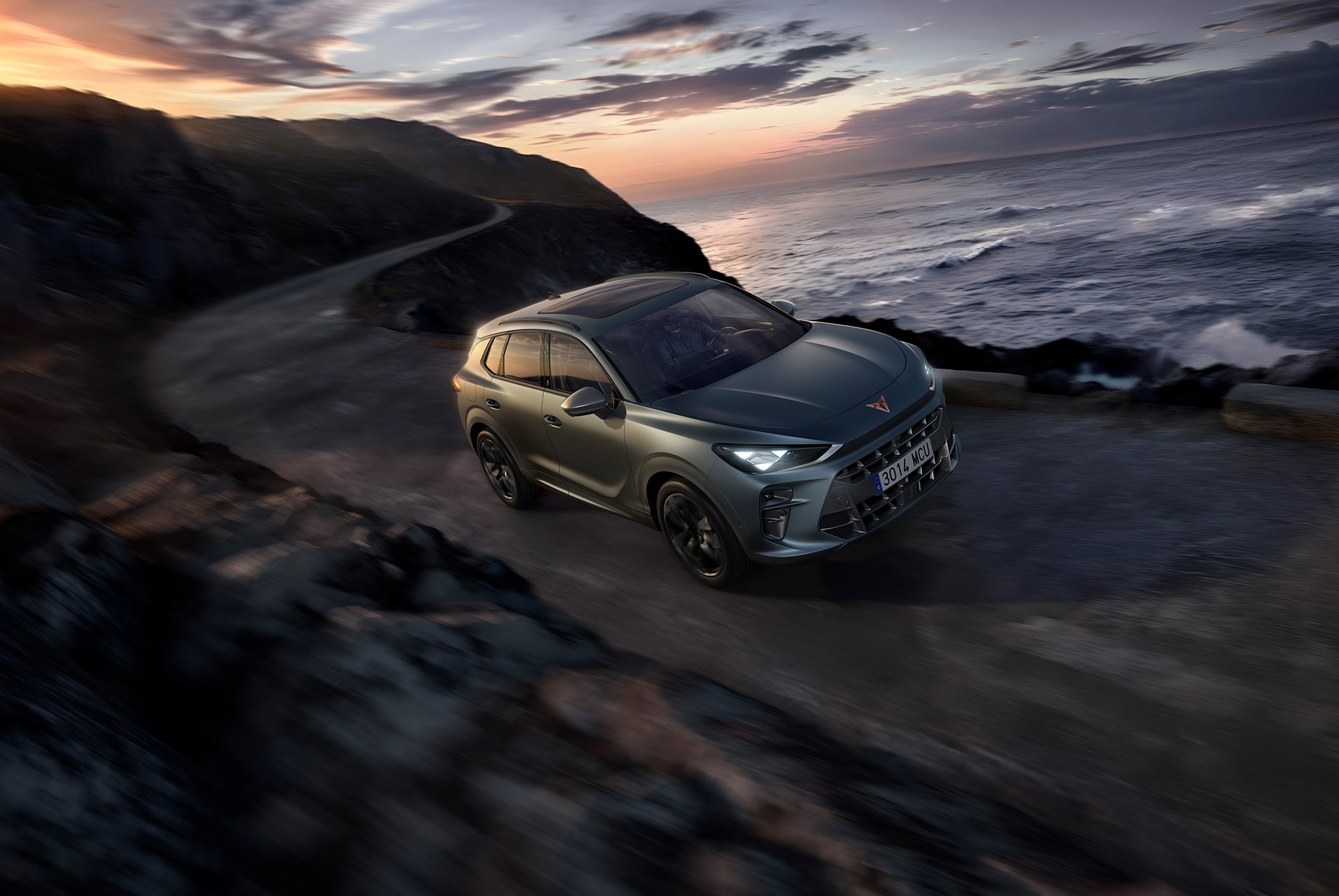 CUPRA Terramar, versió exclusiva America's Cup, gris mat Enceladus, amb llantes d'aliatge de 20 polzades America's Cup, llums de frens CUPRA Matrix LED Ultra HD, i Dynamic Chassis Control (DCC), conduint per una carretera sinuosa mullada vora el mar al capvespre.